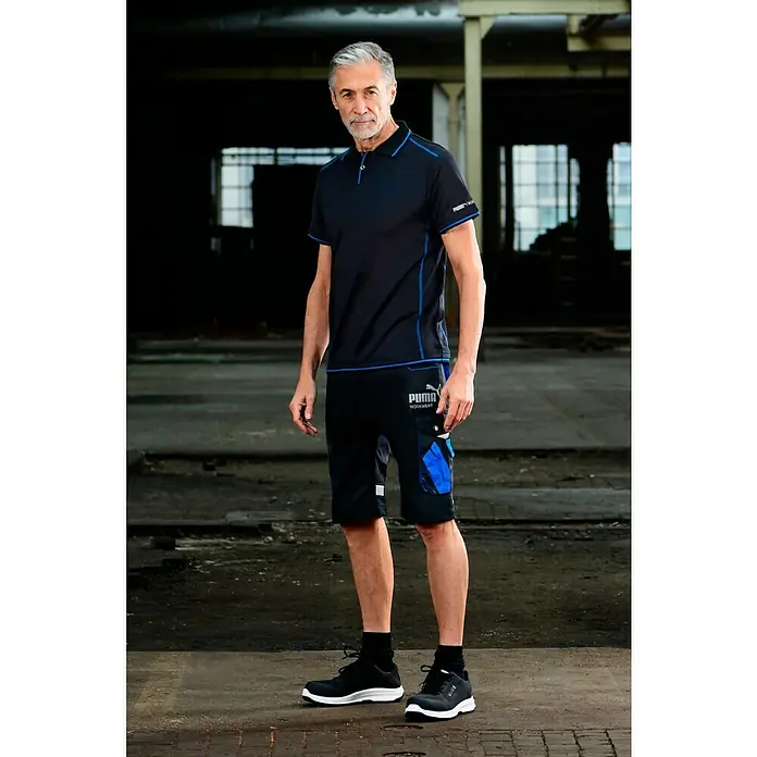 Puma Workwear Poloshirt Champ - Carbon, XL - Mann trägt ein Puma Workwear Outfit, bestehend aus Poloshirt, Shorts und Sicherheitsschuhen, in einer Industrieumgebung.