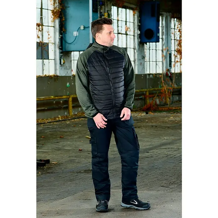 L.Brador Hybridjacke 6090P - Schwarz/Grün, S - Der Mann trägt eine gesteppte Jacke, Arbeitshosen mit Taschen und robuste Stiefel. Geeignet für Arbeit oder Outdoor-Aktivitäten.