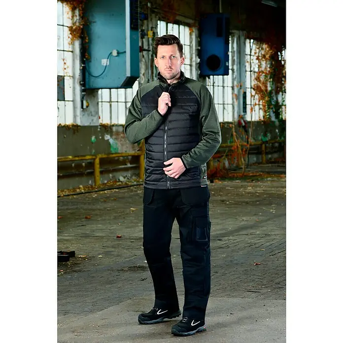 L.Brador Hybridjacke 6090P - Schwarz/Grün, S - Ein Mann in Arbeitskleidung präsentiert eine stilvolle Weste mit gesteppter Vorderseite und praktischen Arbeitshosen mit Taschen.