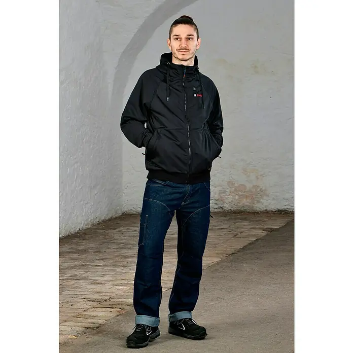 Bosch Professional Arbeitsjacke Hoodie GHH beheizbar - 2 -tlg., Akkuspannung: 12 V, XL - Mann in Bosch beheizbarer Jacke mit Kapuze, blauen Jeans und schwarzen Schuhen vor hellem Hintergrund.