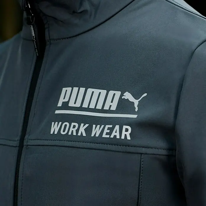 Puma Workwear Softshell-Jacke Champ - Stahlgrau, Carbon, S - Nahaufnahme einer dunkelgrauen PUMA Arbeitsjacke mit weißem Logo und Reißverschluss.