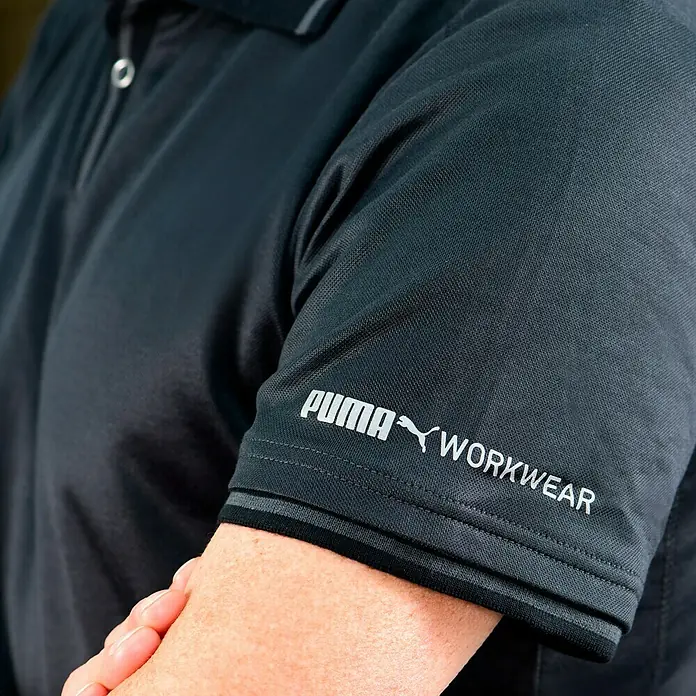 Puma Workwear Poloshirt Champ - Stahlgrau, Carbon, S - Nahaufnahme eines schwarzen Puma Workwear Poloshirts mit weißem Logo am Ärmel.