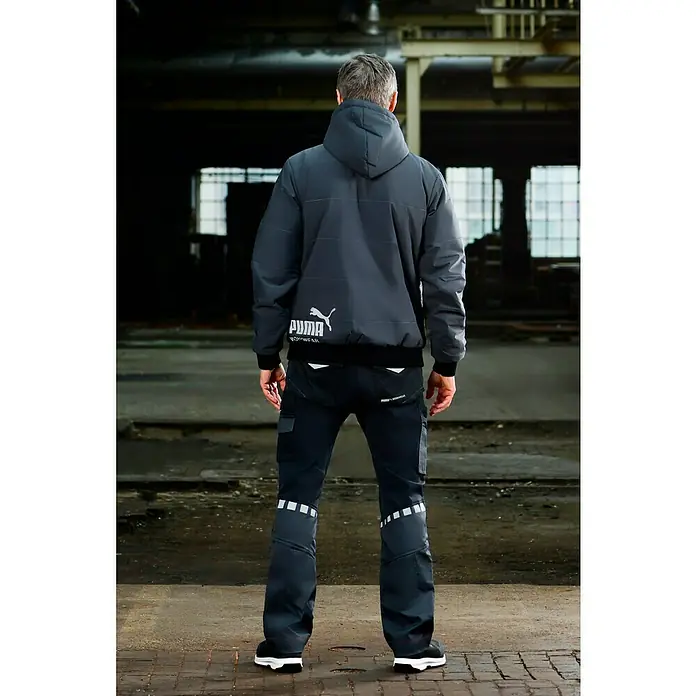 Puma Workwear Winterjacke Champ - Stahlgrau, Carbon, S - Rückansicht eines Mannes in PUMA Workwear Jacke und Hose, stehend in einer Lagerhalle. Die Jacke hat eine Kapuze und das PUMA-Logo.