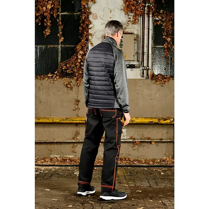 L.Brador Hybridjacke 6090P - Schwarz/Grau, L - Arbeitshose in Schwarz mit orangefarbenen Nähten, kombiniert mit einer ärmellosen Steppjacke in Schwarz und einem grauen Langarmshirt.