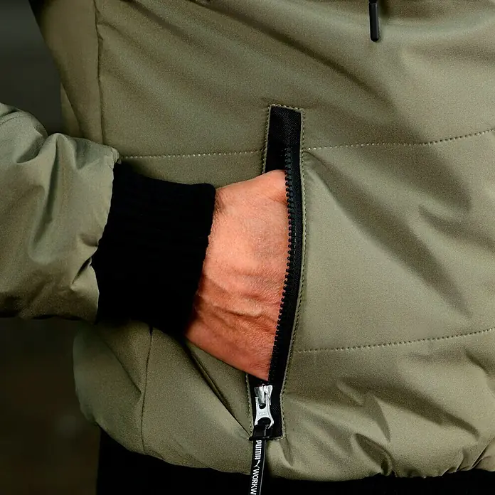 Puma Workwear Winterjacke Champ - Oliv/Carbon, S - Nahaufnahme einer olivgrünen Puma-Arbeitsjacke mit Reißverschlusstasche und einer Hand, die in die Tasche gesteckt wird.