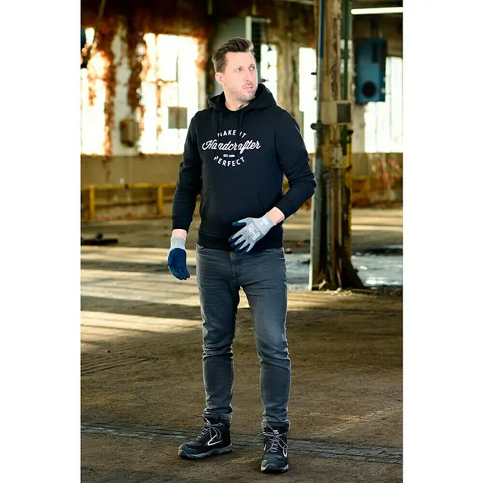 OX-ON Arbeitshandschuhe Recycle Basic 16003 - Grau, 8 - Der Mann trägt eine schwarze Kapuzenjacke mit dem Aufdruck "Make It Handcrafter Perfect", Jeans und Arbeitsstiefel in einer Fabrikhalle.