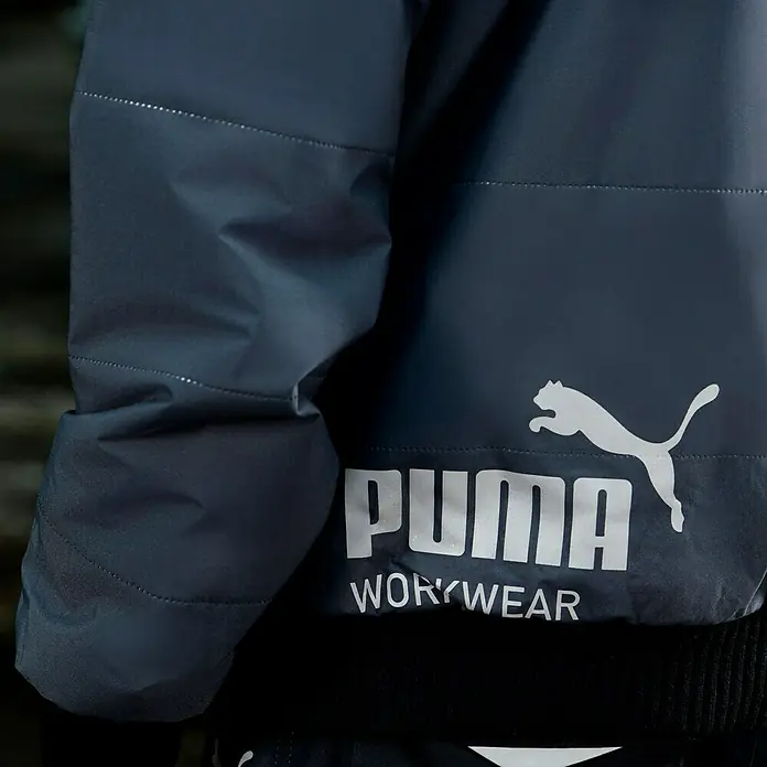 Puma Workwear Winterjacke Champ - Stahlgrau, Carbon, S - Nahaufnahme einer Puma Workwear Jacke in dunklem Blau mit weißem Logo, ideal für den Arbeitsalltag.