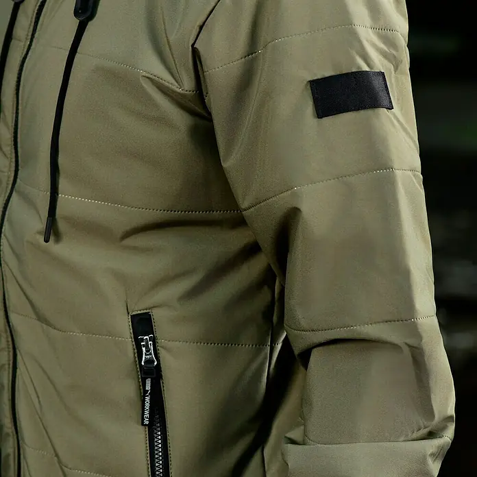 Puma Workwear Winterjacke Champ - Oliv/Carbon, S - Detailaufnahme einer olivgrünen Arbeitsjacke mit Reißverschluss und schwarzem Ärmelpatch. Ideal für den robusten Einsatz im Arbeitsalltag.