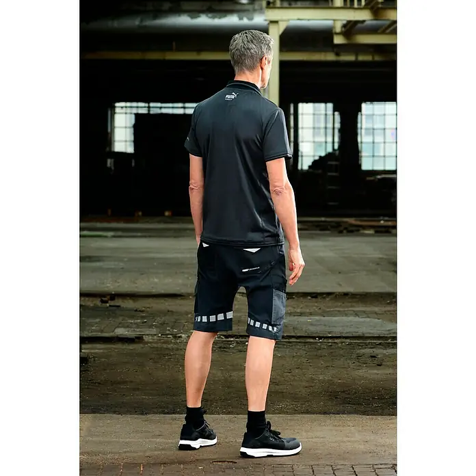 Puma Workwear Poloshirt Champ - Stahlgrau, Carbon, S - Ein Mann von hinten, der Puma Arbeitskleidung trägt, mit einem schwarzen Poloshirt und passenden Shorts mit Reflektoren und schwarzen Schuhen.