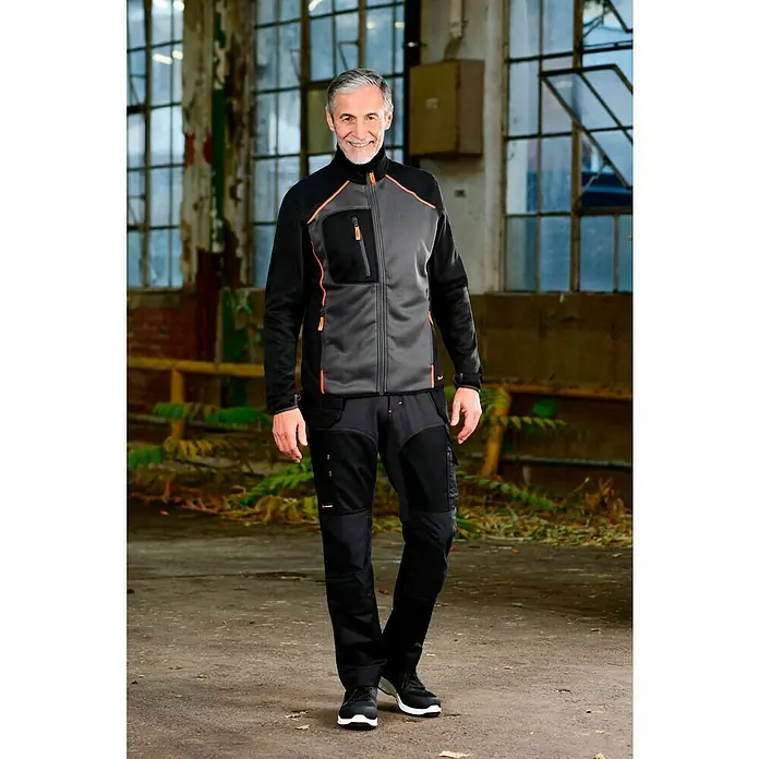 L.Brador Fleecejacke 6195P - Schwarz/Grau, XL - Ein Mann in grauer und schwarzer Arbeitskleidung mit orangen Akzenten. Die Jacke ist geschlossen, die Hose ist schwarz und er trägt schwarze Schuhe.