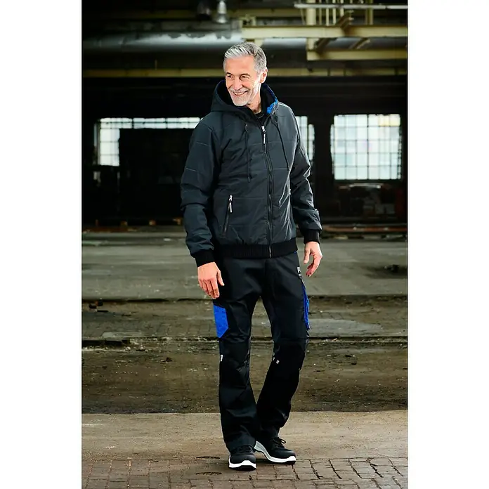 Puma Workwear Winterjacke Champ - Carbon, L - Lachender Mann in schwarzer Jacke und Arbeitshose mit blauen Details. Ideal für Handwerker, die Wert auf Komfort und Funktionalität legen.