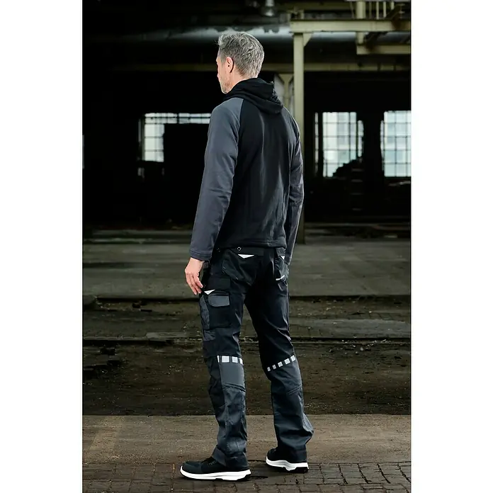 Puma Workwear Kapuzenpullover Champ - Stahlgrau, Carbon, Größe: S - Das Bild zeigt einen Mann von hinten, der eine moderne Arbeitskleidung mit einer schwarzen Kapuzenjacke und Arbeitshosen mit Taschen und Reflektoren trägt.