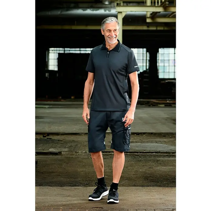Puma Workwear Poloshirt Champ - Stahlgrau, Carbon, S - Ein lächelnder Mann mittleren Alters trägt eine dunkelgraues Puma Polo-Shirt und dunkelgraue Puma Arbeitsshorts und schwarze Sportschuhe.