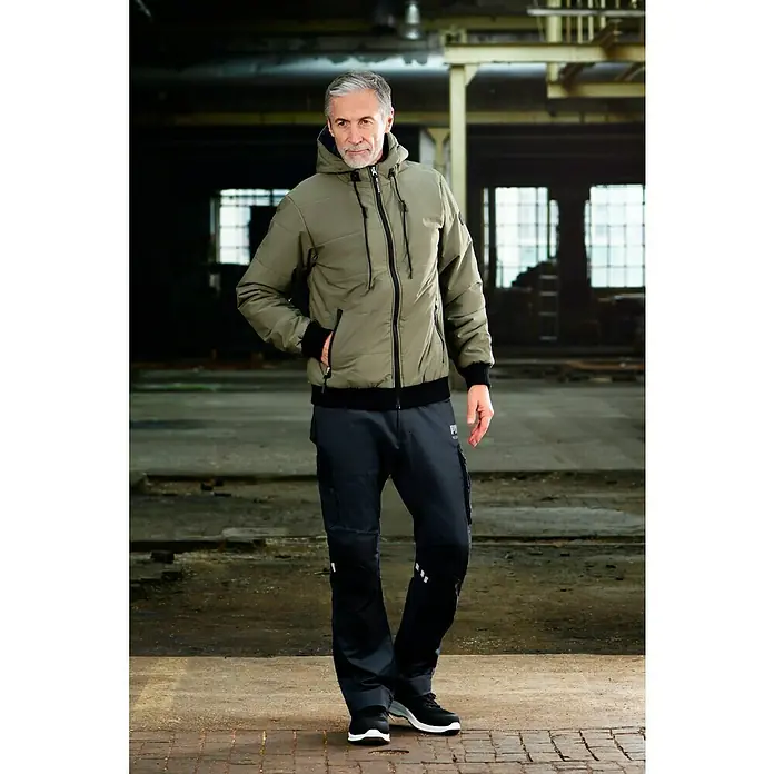 Puma Workwear Winterjacke Champ - Oliv/Carbon, S - Der Mann trägt eine khaki-farbene Jacke mit Kapuze und schwarzem Reißverschluss, kombiniert mit Cargohosen und Sneakern.