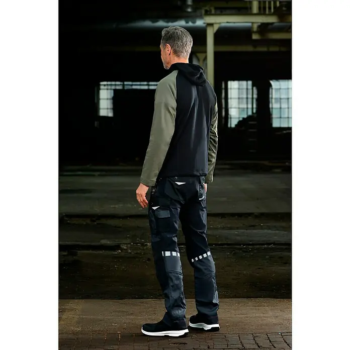 Puma Workwear Kapuzenpullover Champ - Oliv, Carbon, Größe: S - Ein Mann in Arbeitskleidung, bestehend aus einer Kapuzenjacke und Arbeitshosen mit reflektierenden Details, steht in einer Lagerhalle.