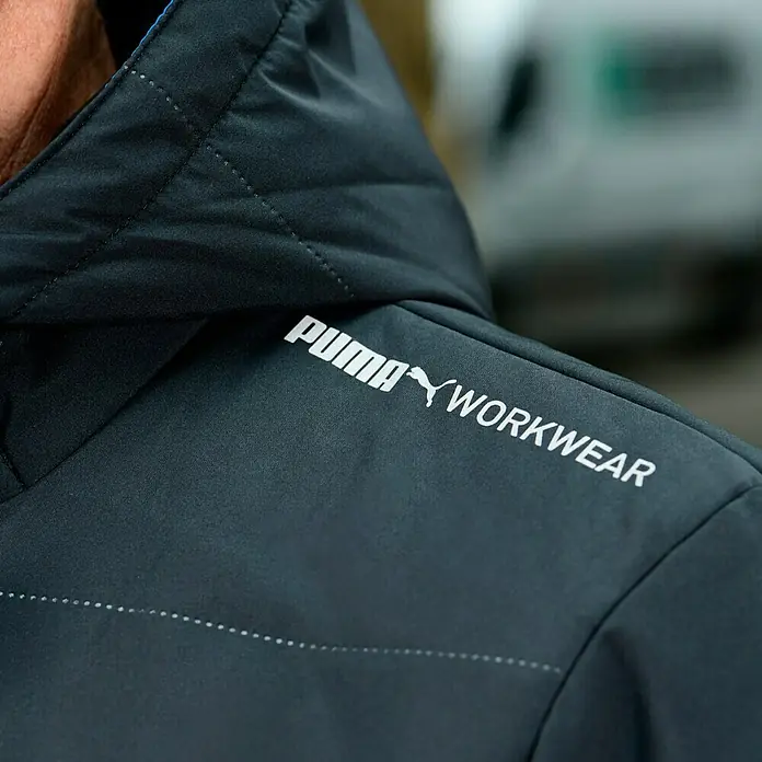Puma Workwear Winterjacke Champ - Carbon, L - Nahaufnahme einer schwarzen Puma-Arbeitsjacke mit dem Puma-Logo und dem Schriftzug "WORKWEAR".
