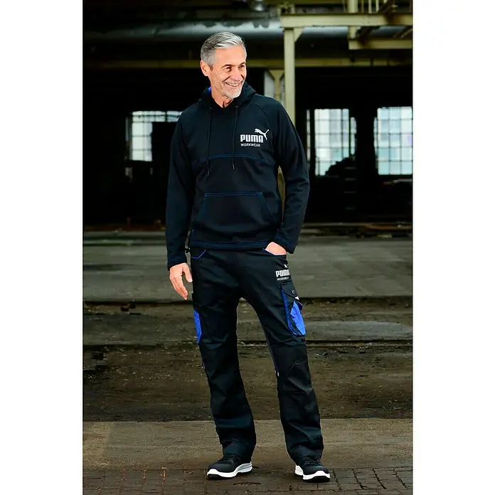 Puma Workwear Kapuzenpullover Champ - Carbon, Größe: XXL - Ein Mann in Puma-Arbeitskleidung, bestehend aus schwarzem Hoodie und passender Hose, posiert selbstbewusst vor Industriehintergrund.
