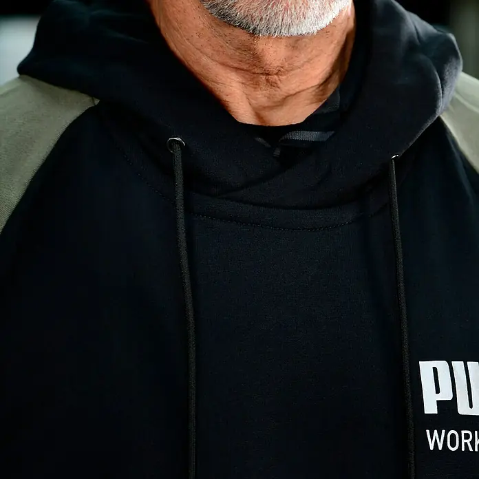 Puma Workwear Kapuzenpullover Champ - Oliv, Carbon, Größe: S - Nahaufnahme eines schwarzen Puma-Hoodies mit Kapuze und Kordelzugdetails, getragen von einem Mann.