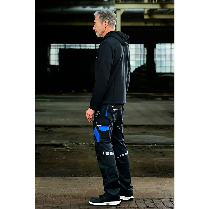 Puma Workwear Kapuzenpullover Champ - Carbon, Größe: XXL - Ein Mann steht in Arbeitskleidung, bestehend aus einer schwarzen Jacke, schwarzen Arbeitshosen mit blauen Akzenten und Sicherheitsschuhen.