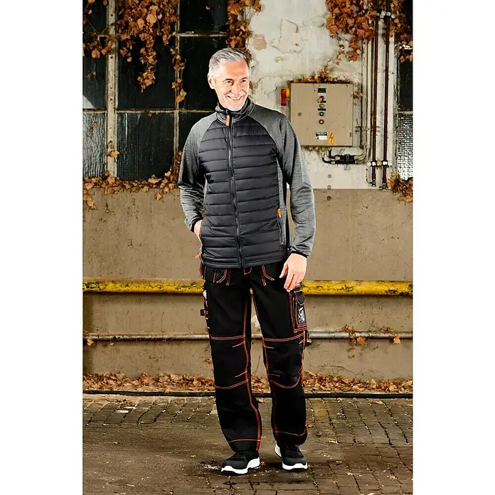 L.Brador Hybridjacke 6090P - Schwarz/Grau, L - Ein Mann in schwarzer Jacke und Arbeitshose steht vor einem Gebäude. Die Jacke ist gesteppt und die Hose hat orangefarbene Details.