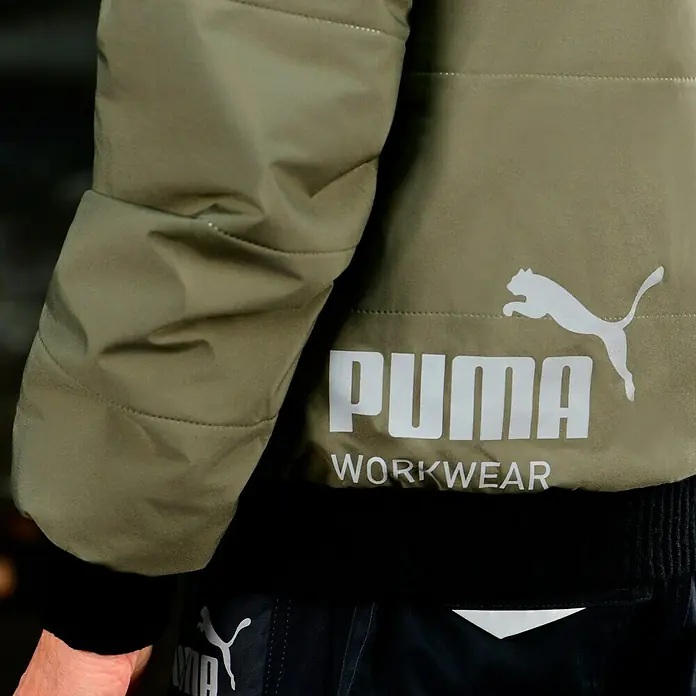 Puma Workwear Winterjacke Champ - Oliv/Carbon, S - Nahaufnahme einer olivgrünen Puma Workwear Jacke mit weißem Logo und Schriftzug, ideal für den Arbeitsalltag.