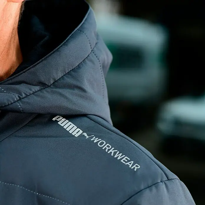 Puma Workwear Winterjacke Champ - Stahlgrau, Carbon, S - Nahaufnahme einer grauen Puma Workwear Jacke mit Kapuze, die das Logo und den Schriftzug gut sichtbar zeigt.