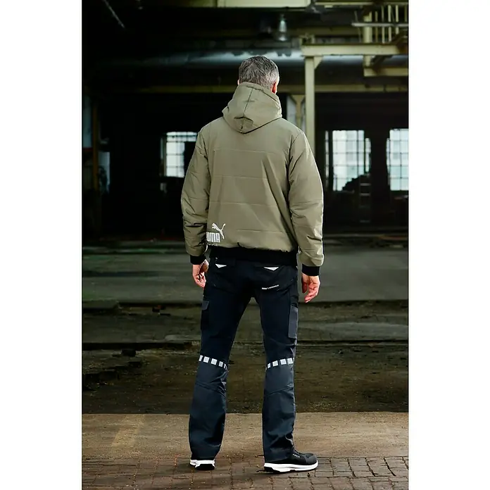 Puma Workwear Winterjacke Champ - Oliv/Carbon, S - Ein Mann von hinten, der eine khaki Puma Jacke mit Kapuze und schwarze Arbeitshosen mit reflektierenden Details und Puma Schuhe trägt.