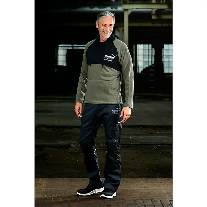 Puma Workwear Kapuzenpullover Champ - Oliv, Carbon, Größe: S - Ein Mann trägt eine Puma Workwear-Hose und einen Hoodie mit Puma-Logo in einer Fabrikhalle. Die Workwear-Kollektion bietet Komfort und Funktionalität.