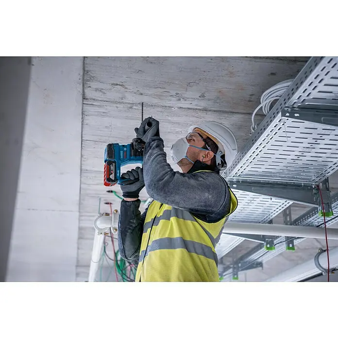 Bosch Professional AMPShare 18V Akku-Bohrhammer PRO GBH 18V-22 - 18 V, 2 Akkus, 4 Ah, 1,9 J - Ein Bauarbeiter bohrt mit einer Bosch-Bohrmaschine ein Loch in eine Betondecke. Er trägt Schutzkleidung wie Helm und Maske.