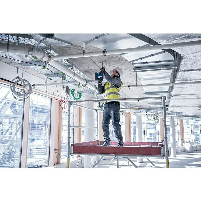 Bosch Professional AMPShare 18V Akku-Bohrhammer PRO GBH 18V-22 - 18 V, 2 Akkus, 4 Ah, 1,9 J - Ein Bauarbeiter auf einem Gerüst montiert mit einer Bohrmaschine Rohre an der Decke eines Rohbaus, wobei die Rohre und die Baustelle im Fokus stehen.