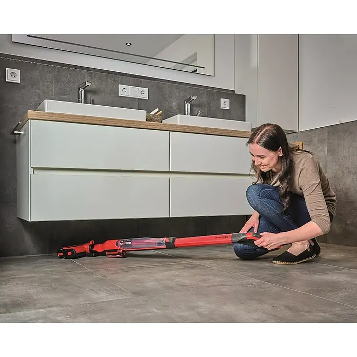 Einhell Power X-Change 18V Akku-Hartbodenreiniger Cleanexxo - 18 V, Ohne Akku - Frau reinigt mit dem roten Einhell Akku-Bodenreiniger den Boden unter dem Waschtisch im Badezimmer.