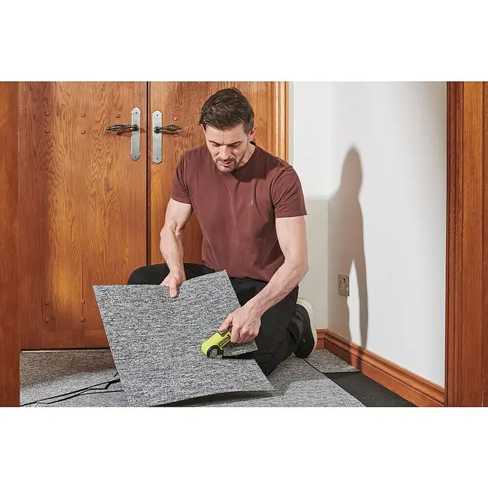 Ryobi 4V USB Akku-Universalschere RCT4-120G - 4 V, 1 Akku, 2 Ah - Ein Mann schneidet eine graue Teppichfliese mit einem gelben Werkzeug. Es sieht so aus, als würde er den Boden mit Teppichfliesen auslegen.