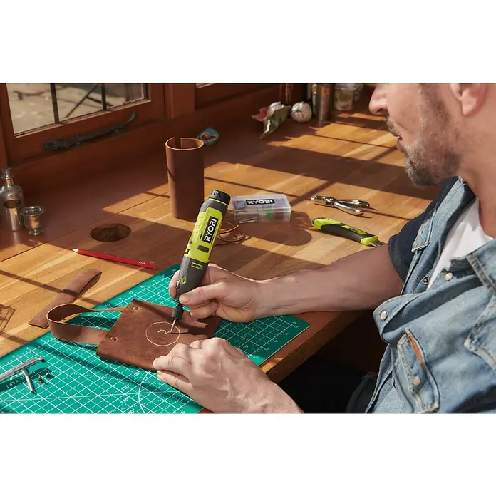 Ryobi 4V USB Akku-Multifunktionswerkzeug RRT4-120GA15 - 4 V, 1 Akku, 2 Ah - Nahaufnahme eines Mannes, der mit einem gelben Ryobi-Rotationswerkzeug Leder bearbeitet, platziert auf einer grünen Schneidematte auf einem Holztisch.