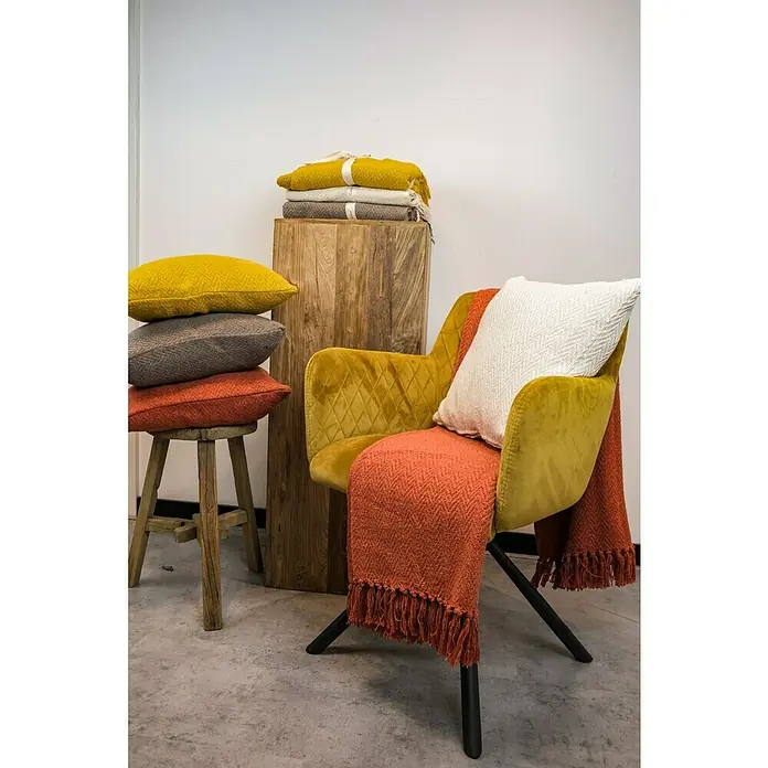 Decke Kent - Golden Yellow, 170 x 130 cm, 100 % Baumwolle - Gemütliche Szene mit einem senfgelben Sessel, orangefarbener Decke und Kissen auf einem Holzhocker.