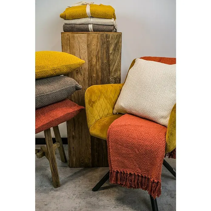 Decke Kent - Golden Yellow, 170 x 130 cm, 100 % Baumwolle - Dekorative Kissen und Decken in verschiedenen Farben auf einem Holzstuhl und einem Sessel arrangiert.