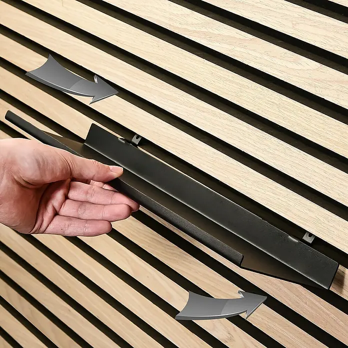 Millex Regal XL für Akustikpaneel - L x B x H: 10 x 40 x 2,5 cm, Passend für: Akustikpaneele, Montageart: Einhängen - Nahaufnahme einer Hand, die ein schwarzes Metallregal an einer Holzlattenwand befestigt. Das Regal ist schlank und modern.