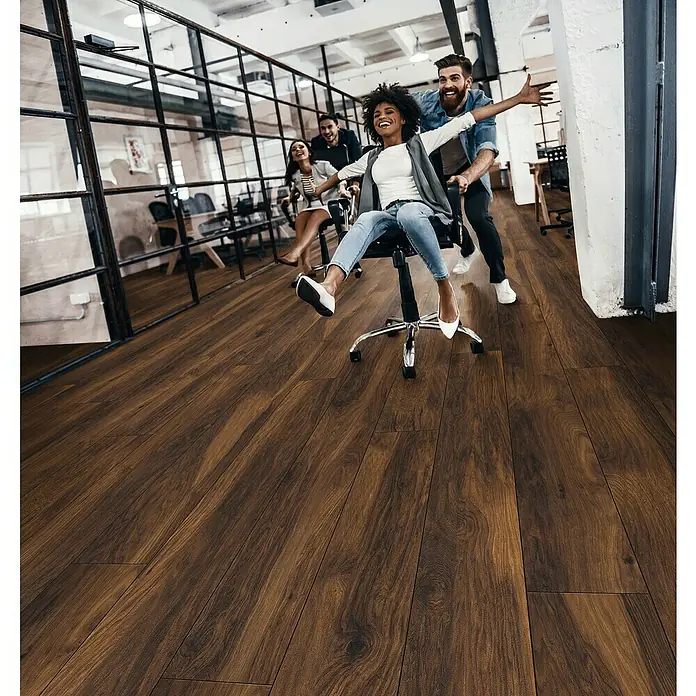 Krono Original Laminatmuster Atlantic 10 Red River Hickory - 297 x 195 x 10 mm, Landhausdiele, Red River Hickory - Büroszene mit elegantem Parkettboden: Eine Gruppe junger Leute hat Spaß bei einem Bürostuhlrennen auf dem hochwertigen Holzboden.