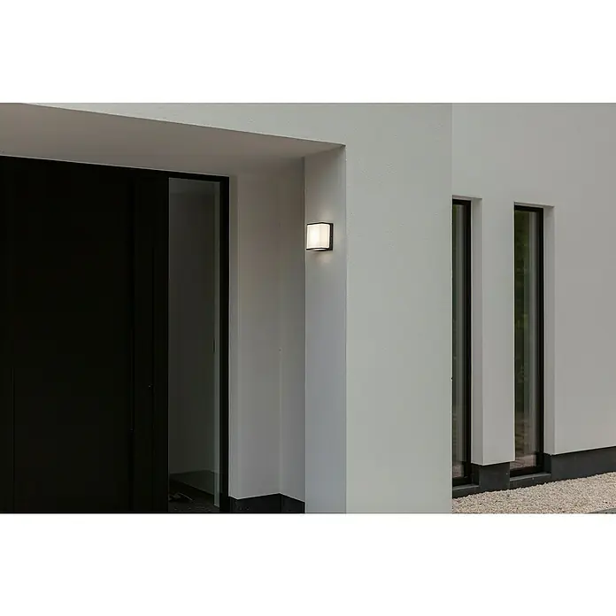 Lutec Doblo Aplique exterior LED Flat - 9 W, 8 x 15 x 15 cm, Negro mate, IP54 - Este primer plano muestra una lámpara de pared exterior moderna que proyecta un brillo cálido sobre una pared blanca, junto a una puerta oscura.