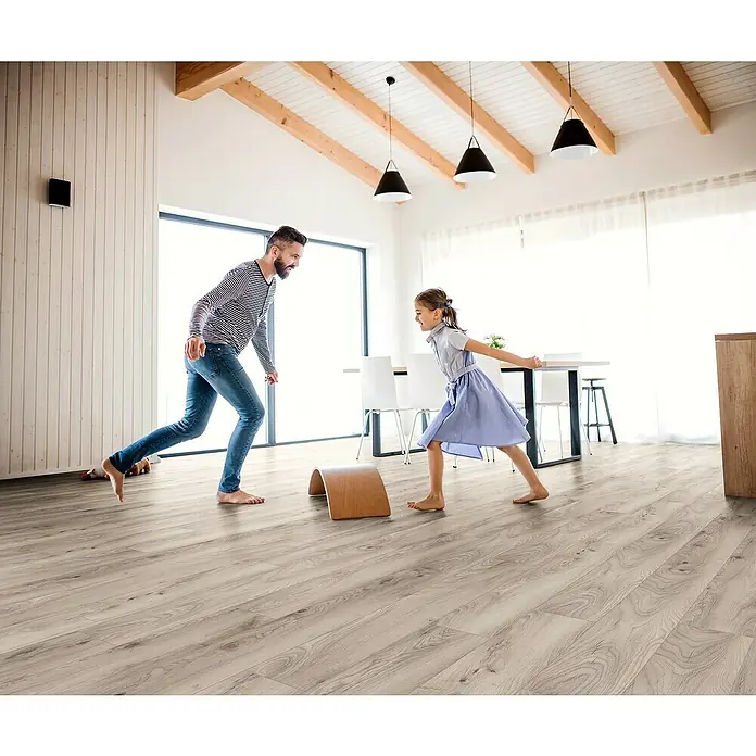 Krono Original Laminat Organic Flex - Pebble Manor Oak, Landhausdiele, 1.288 x 189 x 7 mm - Heller Holzfußboden in einem modernen Haus, wo ein Vater und eine Tochter barfuß spielen, vermittelt ein Gefühl von Wärme und Behaglichkeit.