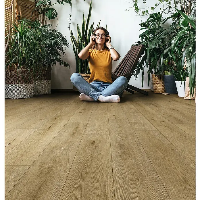 Krono Original Laminatmuster Organic Flex Flax River Oak - 297 x 189 x 7 mm, Landhausdiele - Frau sitzt mit Kopfhörern auf einem Holzboden umgeben von Pflanzen. Der Holzboden ist hell und weist eine natürliche Maserung auf.