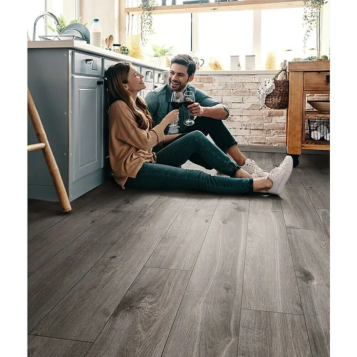 Krono Original Laminat Organic Classic - Tornado Oak, Landhausdiele, 1.288 x 195 x 8 mm - Helles, graues Laminat mit Holzstruktur verlegt, im Hintergrund ein Paar beim Wein trinken. Modernes und stilvolles Bodendesign.