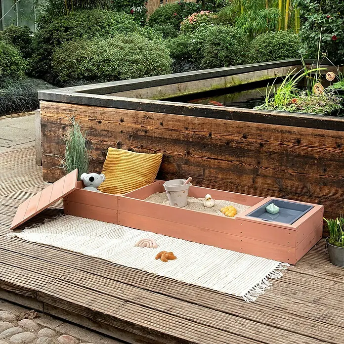 Muddy Buddy Sandkasten Space Saver - 150 x 45 x 20 cm, Holz, Puderrosa - Ein sandfarbener Sandkasten mit Spielzeug und einer Wasserschale steht auf einem Teppich auf einer Holzterrasse im Garten.