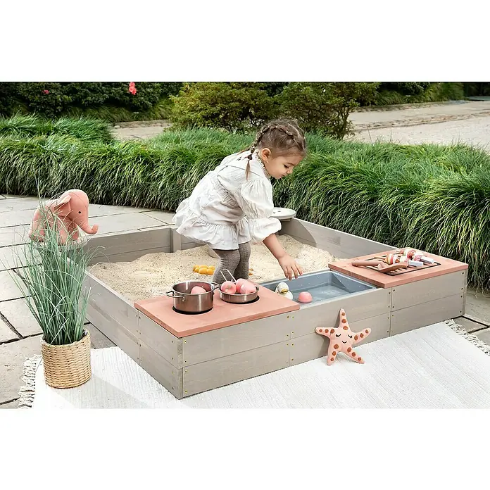 Muddy Buddy Sandkasten Wild Bear - 120 x 120 x 20 cm, Holz, Warmgrau/ Puderrosa - Ein Kind spielt im Outdoor-Spielcenter mit Sandkasten und Wasserspiel, ergänzt durch Spielzeug wie einen Plüschelefanten und Kochtöpfe.