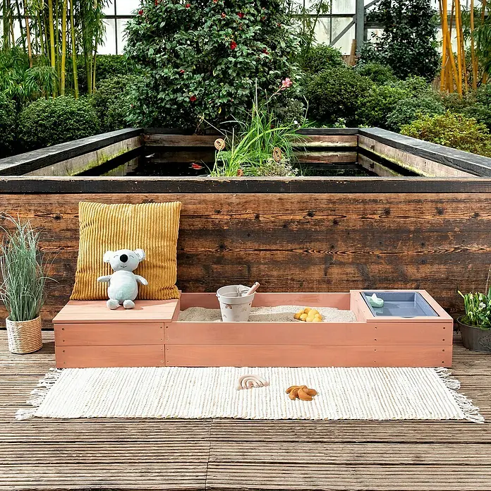 Muddy Buddy Sandkasten Space Saver - 150 x 45 x 20 cm, Holz, Puderrosa - Ein Kinder Sand- und Wassertisch aus Holz mit Spielzeug auf einer Terrasse mit einem Kissen und einem Koala Bär.
