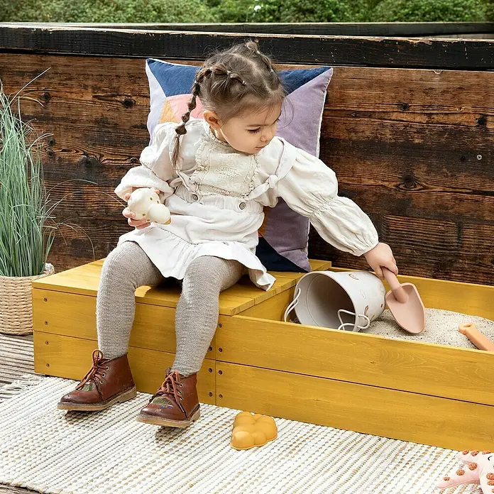 Muddy Buddy Sandkasten Space Saver - 150 x 45 x 20 cm, Holz, Ocker - Ein kleines Mädchen sitzt auf einer gelben Sandkiste mit Spielzeug. Sie trägt ein helles Kleid, Strumpfhosen und braune Schuhe.
