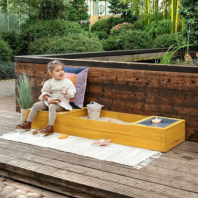 Muddy Buddy Sandkasten Space Saver - 150 x 45 x 20 cm, Holz, Ocker - Ein sonnengelber Sandkasten mit Wasserspielbereich und einem kleinen Mädchen, das darauf sitzt. Perfekt für kreatives Spielen im Freien.