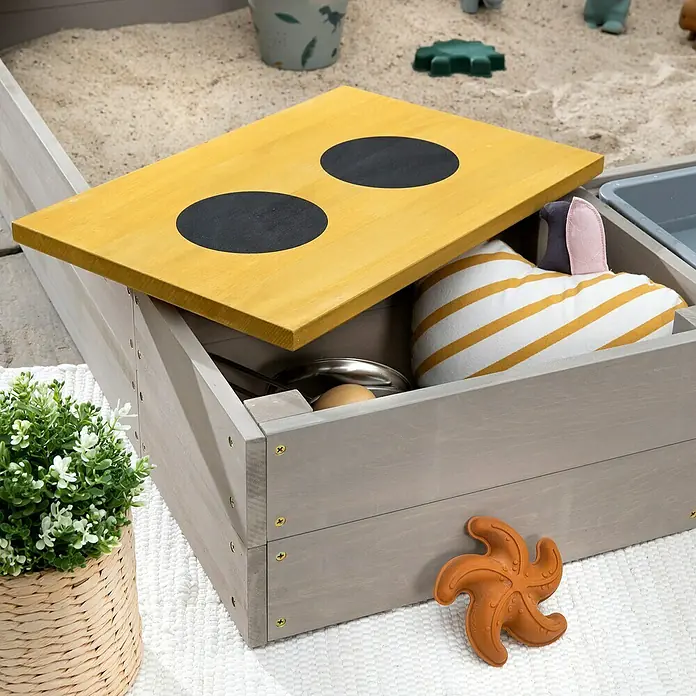 Muddy Buddy Sandkasten Wild Cat - 120 x 120 x 20 cm, Holz, Warmgrau/ Ocker - Nahaufnahme einer grauen Spielzeugkiste mit gelbem Deckel, gefüllt mit Spielzeug auf einem weißen Teppich. Eine Pflanze steht links.