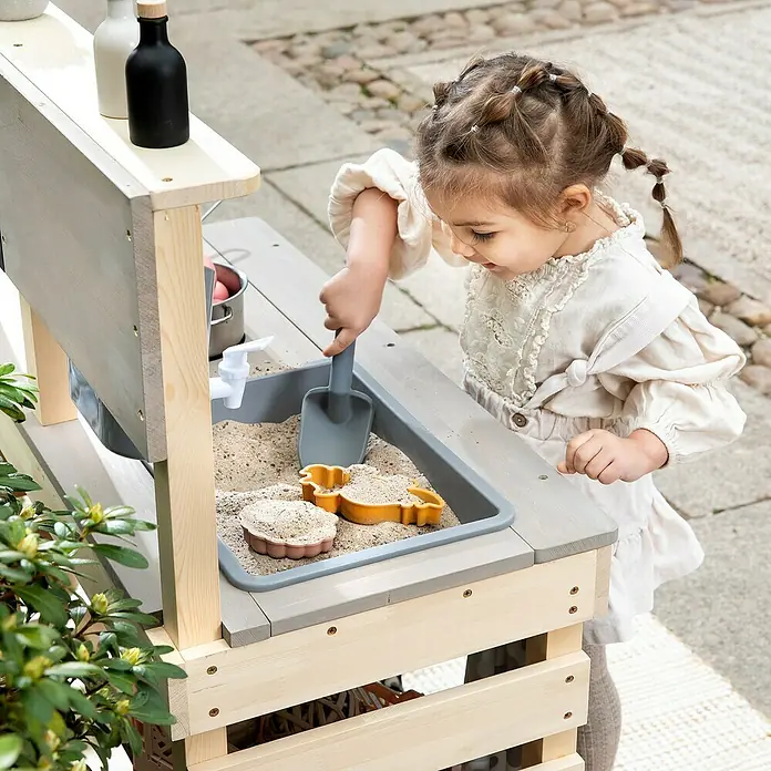 Muddy Buddy Matschküche Big Leo - 80 x 40 x 90 cm, Holz, Natur/ Grau - Ein kleines Mädchen spielt mit einem Outdoor-Spielküchen-Set, das eine Spüle mit Sand und Formen enthält.