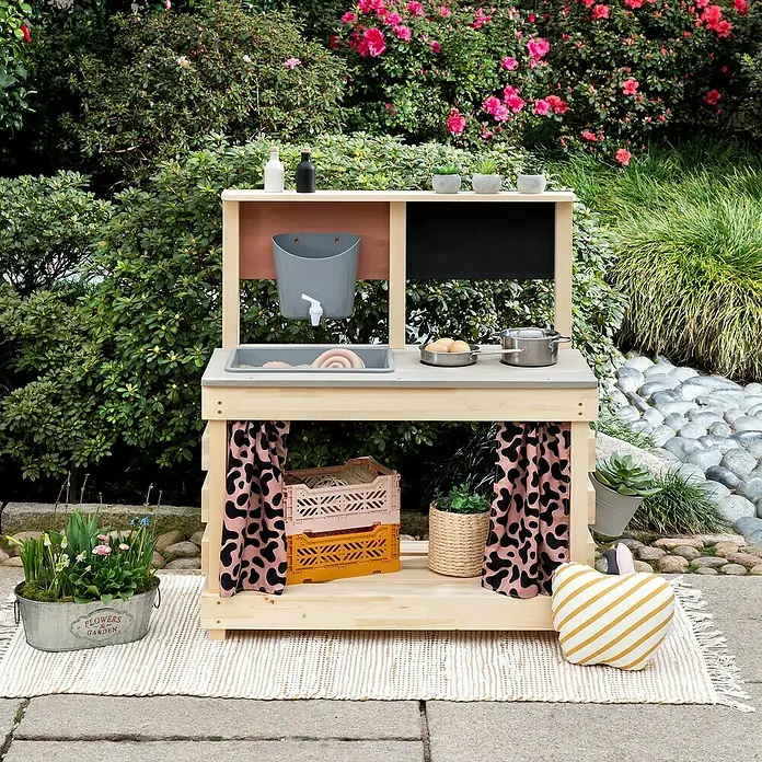Muddy Buddy Matschküche Big Leo - 80 x 40 x 90 cm, Holz, Natur/ Puderrosa - Ein hölzerner Outdoor-Spielküche mit Wasserhahn, Spüle und Stauraum. Die Küche ist mit Accessoires geschmückt und steht im Garten.