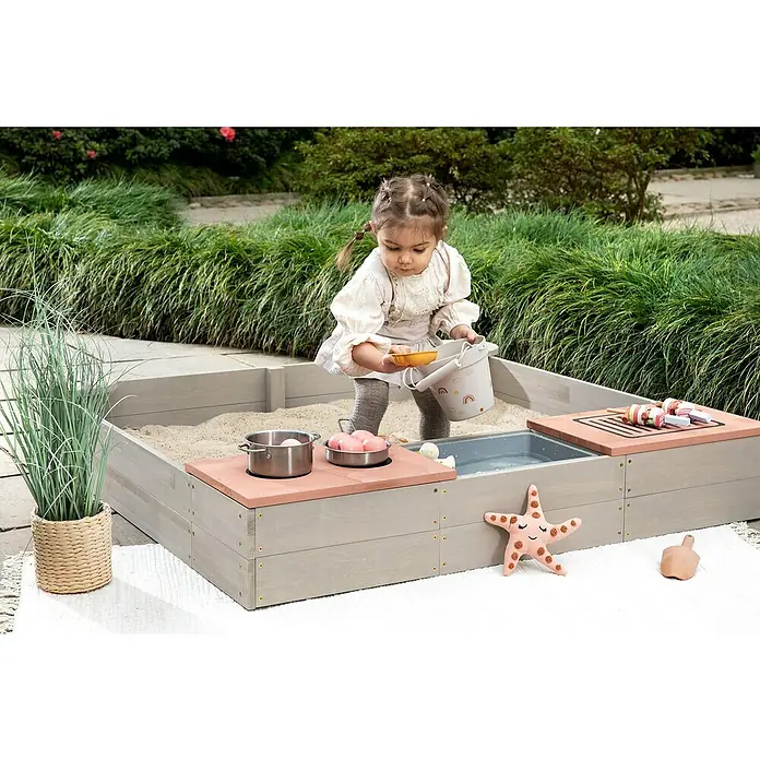 Muddy Buddy Sandkasten Wild Bear - 120 x 120 x 20 cm, Holz, Warmgrau/ Puderrosa - Ein kleines Mädchen spielt in einem grauen Sandkasten mit Spielküche und Zubehör im Garten.